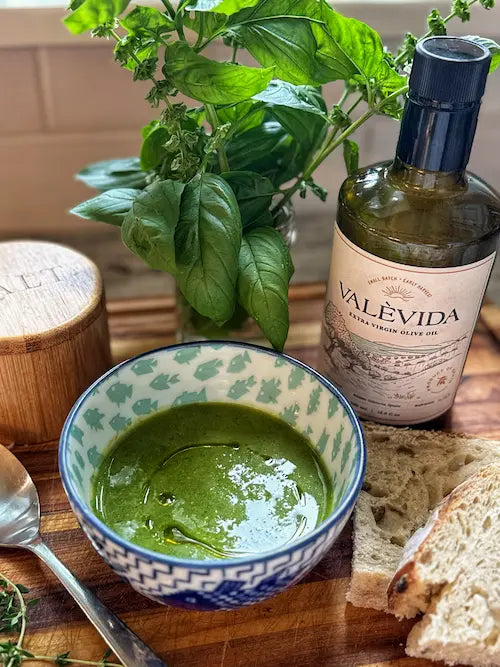 Bowl of green broccoli soup drizzled with Valèvida extra virgin olive oil, next to the bottle and sourdough bread