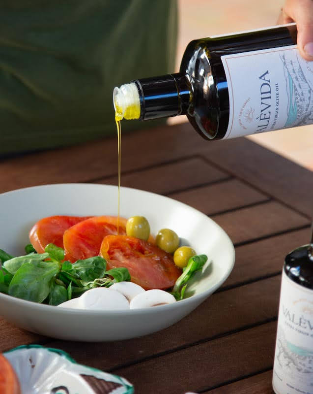 A hand holds a bottle of VAlevida olive oil, pouring a stream of golden oil into a white bowl of salad containing tomatoes, olives, and greens, set on a dark wooden table.