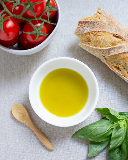 A white bowl filled with Valevida extra virgin olive oil on a light linen surface, surrounded by a wooden spoon, fresh basil leaves, a sliced baguette, and a bowl of red cherry tomatoes.