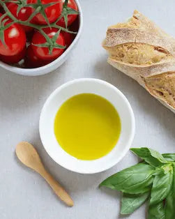 A white bowl filled with Valevida extra virgin olive oil on a light linen surface, surrounded by a wooden spoon, fresh basil leaves, a sliced baguette, and a bowl of red cherry tomatoes.