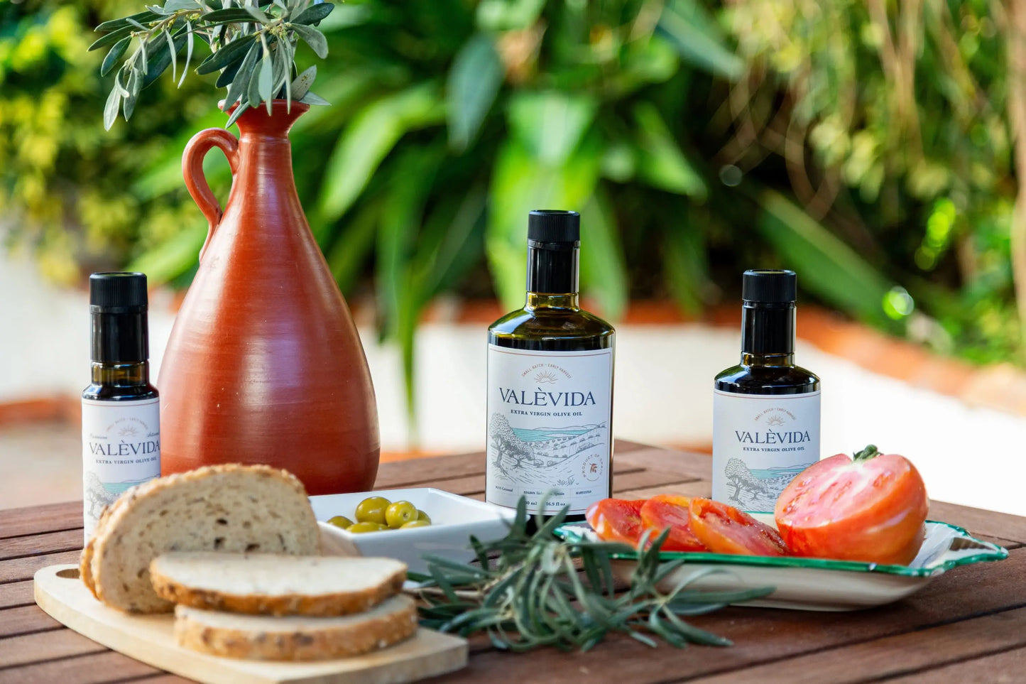 Valevida olive oil bottles with bread, tomatoes, and olives on a wooden table outdoors.