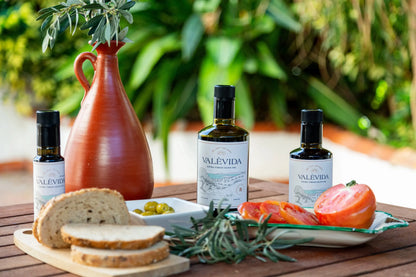 Valevida olive oil bottles with bread, tomatoes, and olives on a wooden table outdoors.